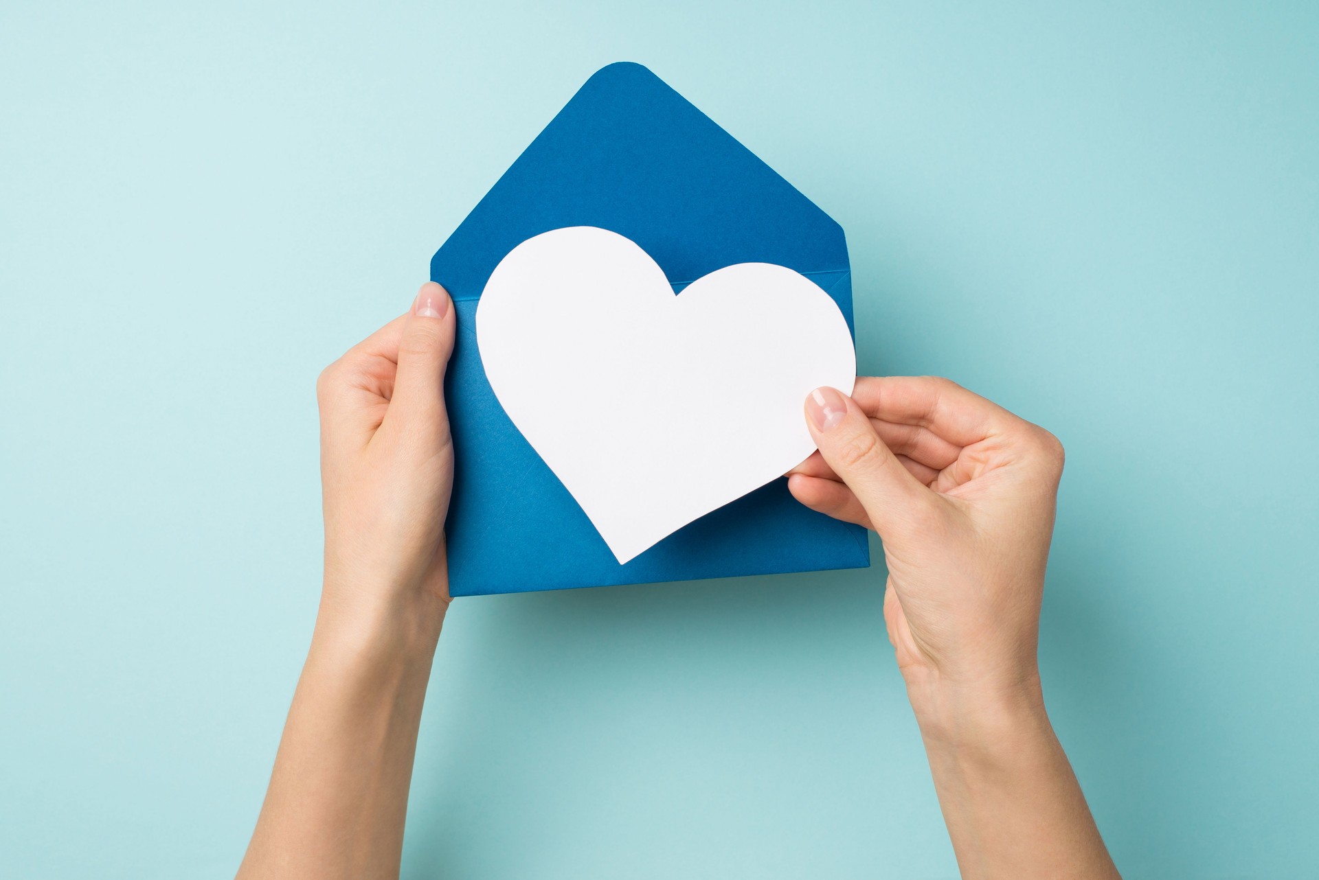 First person top view photo of female hands holding open blue envelope with white paper heart on isolated pastel blue background with blank space